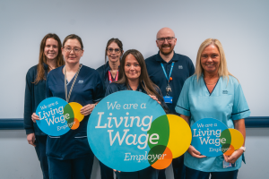 Group of six NHS 24 employees. They are all wearing different NHS Scotland uniforms and are smiling to the camera. Three people at the front of the photo are each holding a Living Wage logo board which has "We are a living wage employer" printed on it. 
