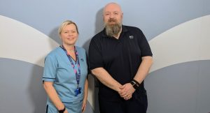 Photo of Tracy and David standing together and smiling. Tracy is in a light blue NHS Scotland tunic. David is wearing a navy blue NHS Scotland polo shirt.