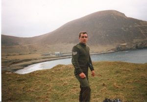 A person in military uniform stands on a grassy hill overlooking a calm bay with a large, rocky hill in the background. The sky is overcast, and the landscape has a rugged, remote feel to it.