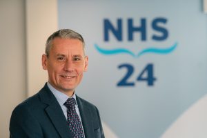 Photograph of Patrick Rafferty, Director of Nursing and Care. He stands in front of the NHS 24 logo.