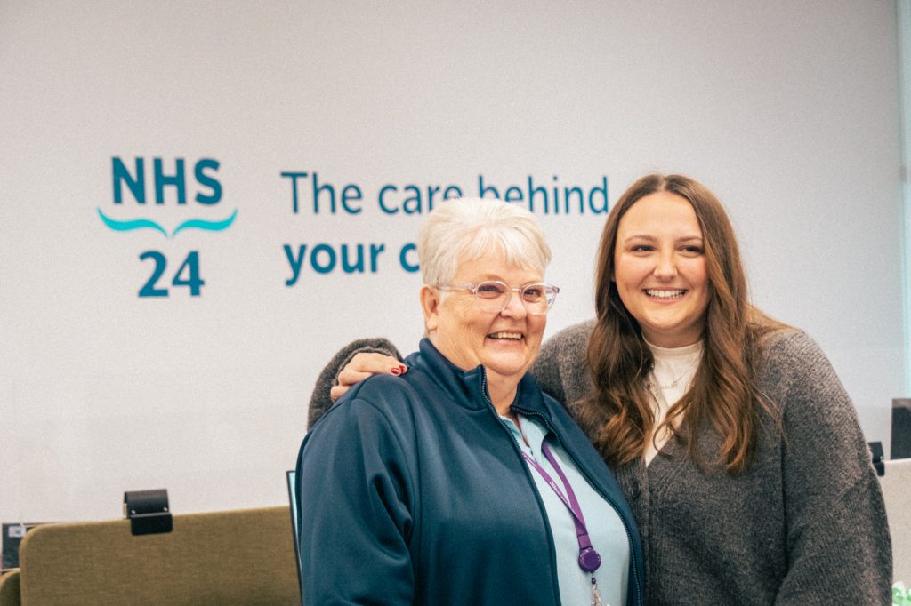 Photo of NHS 24 Call Handler Shirley and patient Daisy. Shirley has short white hair, and is wearing glasses and her NHS uniform. Daisy has her arm around Shirley's shoulder and has long dark hair. Both are smiling widely. 