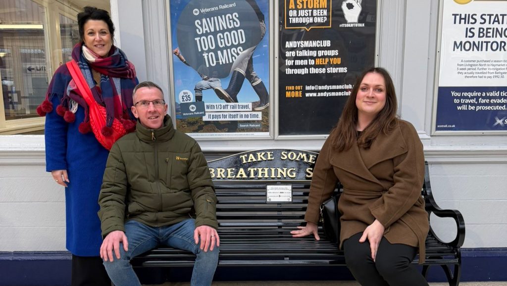Three people are posed around a black bench with the inscription "Take Some Breathing Space." The person on the left stands in a blue coat and colourful scarf, the person in the middle sits wearing a green jacket and jeans, and the person on the right is sitting in a brown coat. Behind them, posters with various messages are visible.